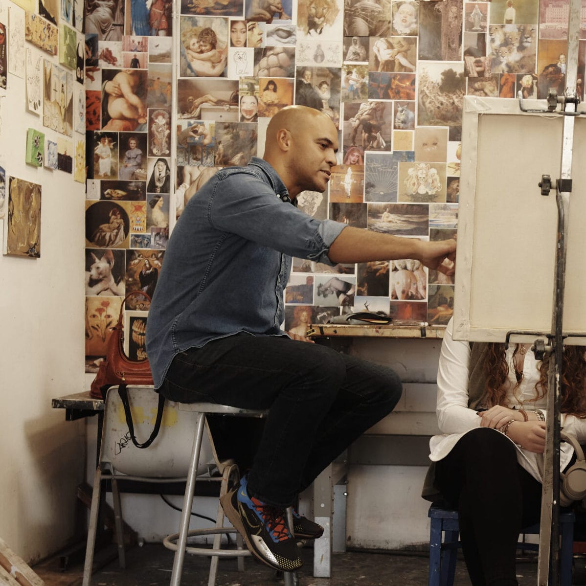 A faculty member sits on a stool in a student's painting studio, offering feedback on their work.