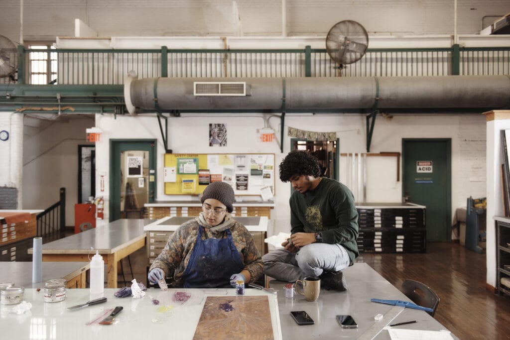 Two people in an art studio at MassArt are engrossed in creative projects. One is seated, painting, while the other, holding a cup, looks on. The space boasts art supplies, paintings on tables, and industrial design elements like exposed ducts and fans—a vibrant hub of education and creativity.