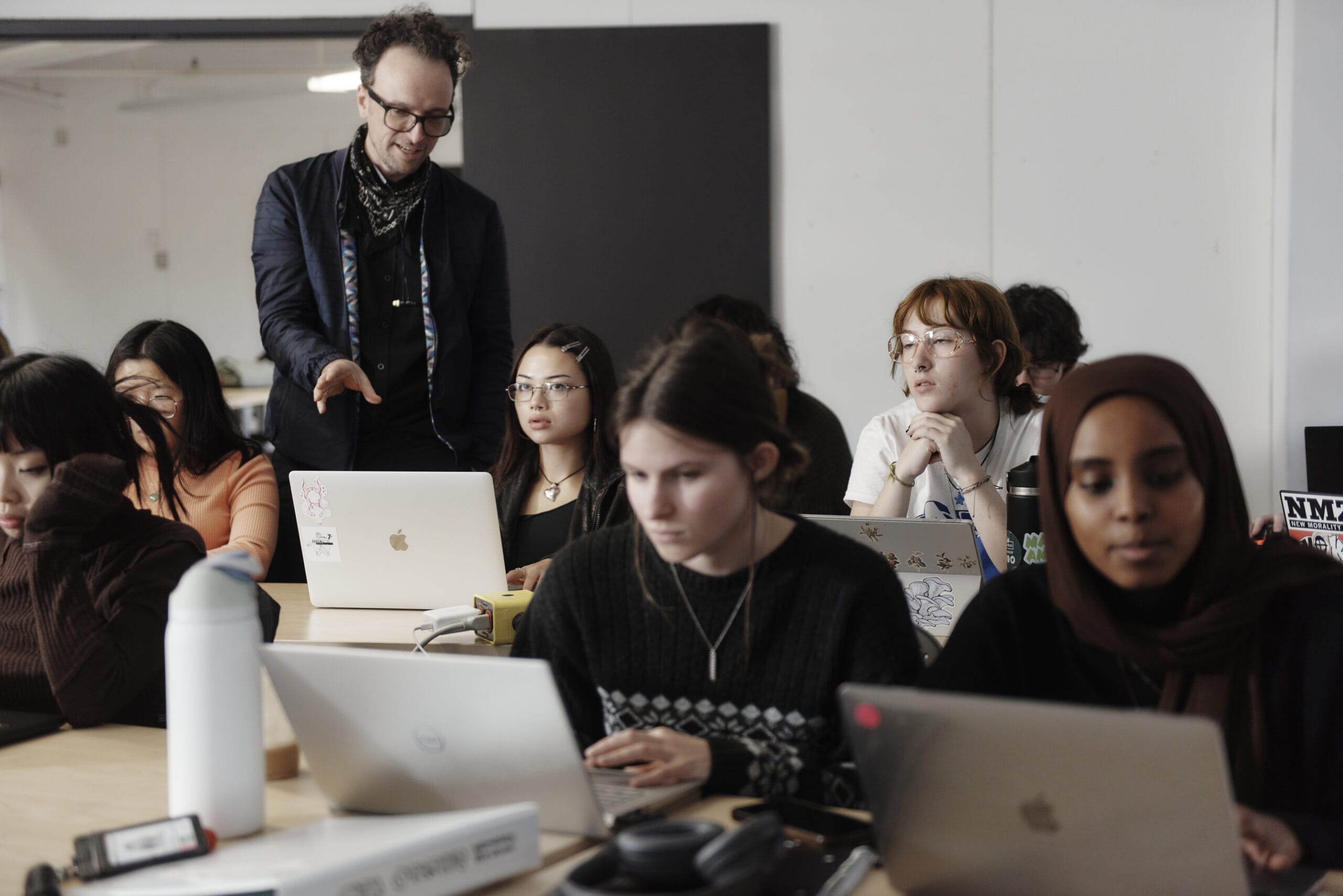 A professor stands amidst a classroom full of students on laptops and points to a laptop as he speaks to a student.
