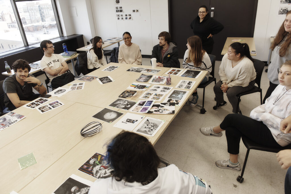 A diverse group of people, representing various genders and ethnicities, are seated around large tables in a room filled with printed images and papers. Engaged participants in the Communication Design program are either examining the visuals or interacting with each other.