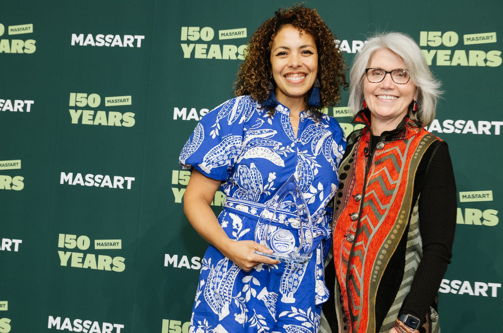 Two women stand smiling with an award in front of a green backdrop that says MassArt 150 years.