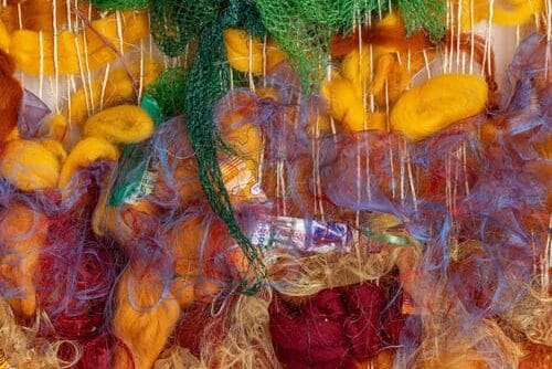 Colorful fibers in red, orange, and yellow intertwine with green netting and candy wrappers, echoing the vibrant creativity often seen at MFA events or showcased by artists in low residency programs, all layered in a textured, abstract arrangement.
