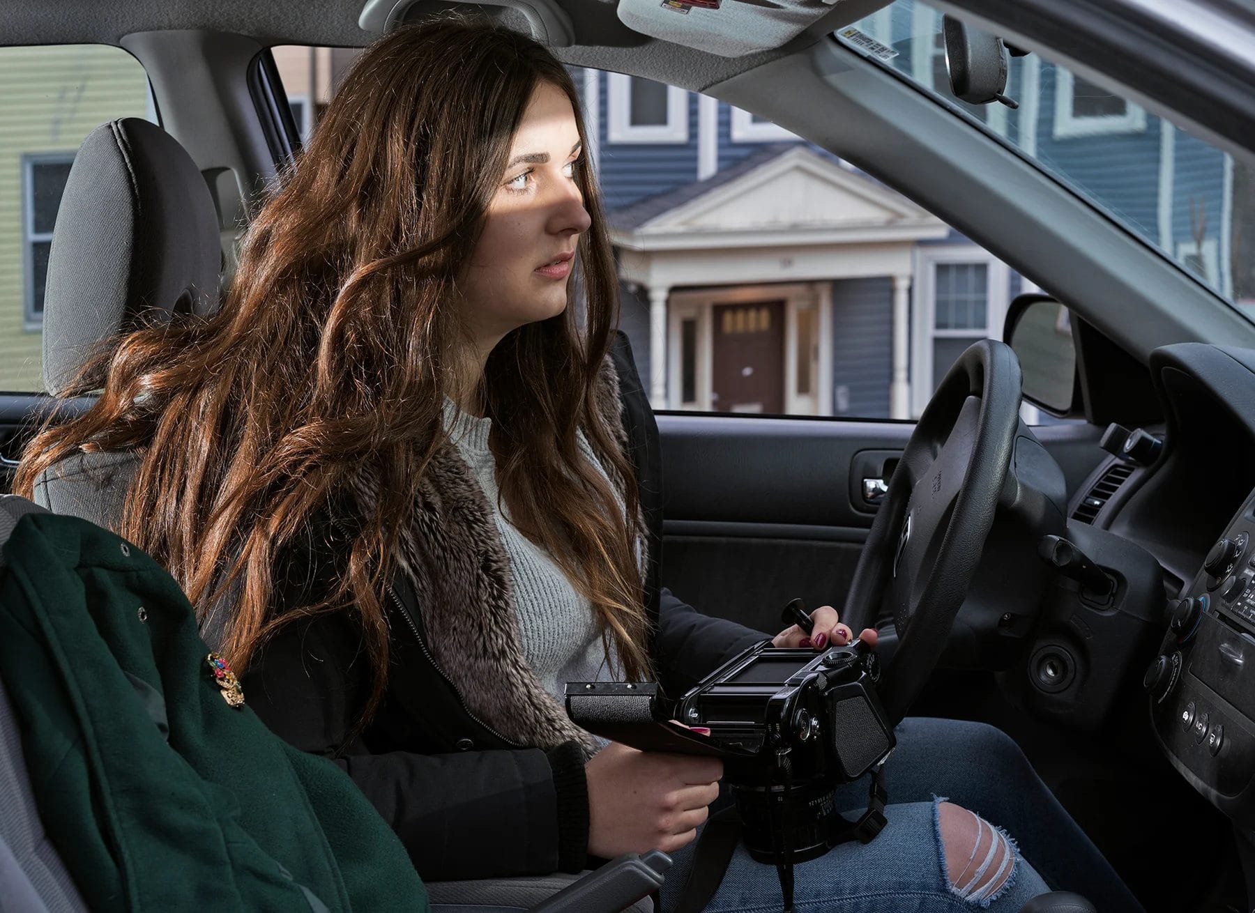 A young woman with long brown hair, pursuing a Photography BFA, sits in the driver’s seat of a car, holding a video camera and looking out the window toward colorful houses. A green jacket is draped over the passenger seat.