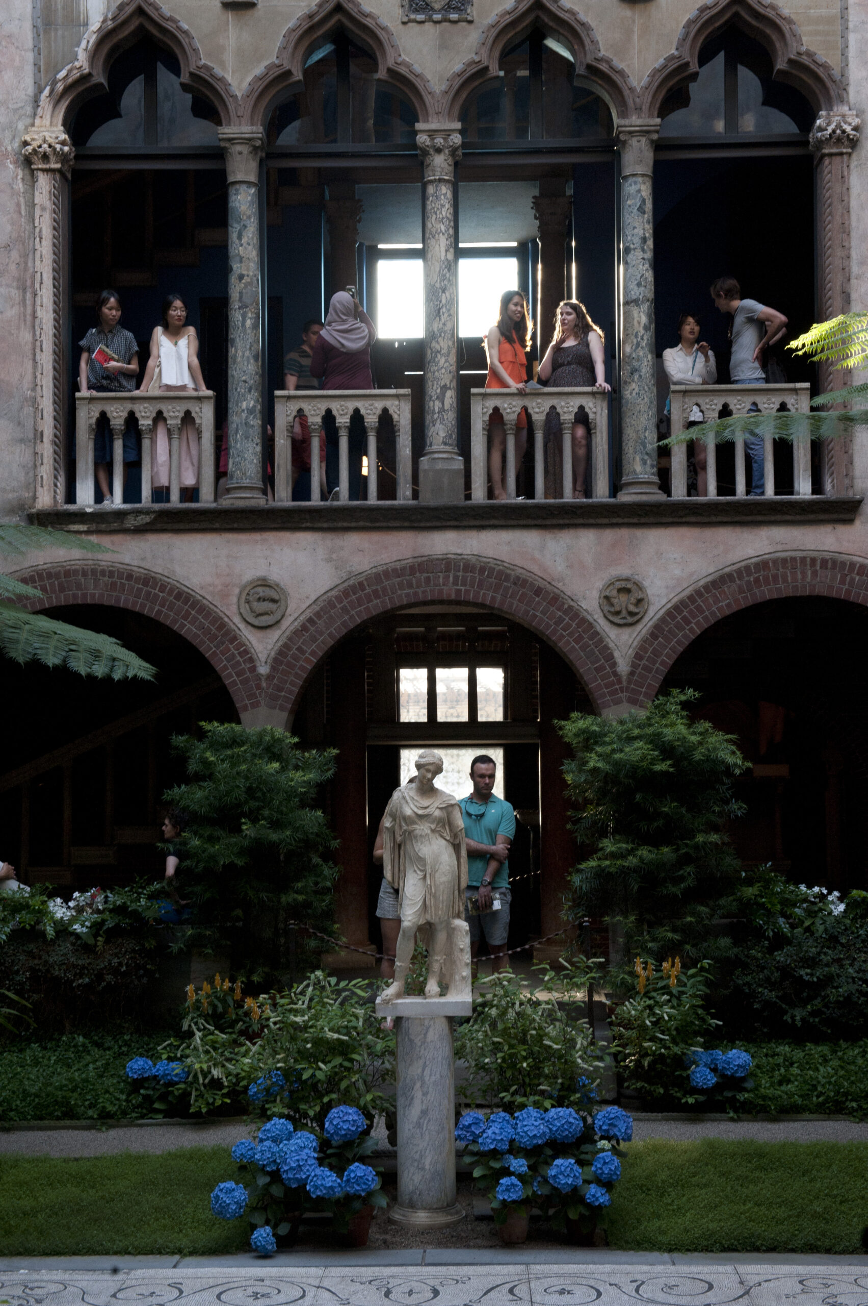 The courtyard of the Isabella Gardner Museum showing a statue at the bottom of the photo and a back-lit balcony with people gathered and talking looking down upon the courtyard.