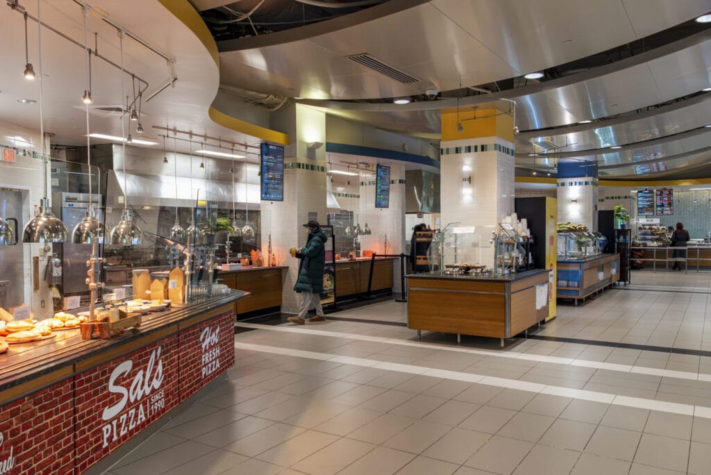 A spacious food court offers diverse dining options, including Sals Pizzeria. A few people are browsing amid the sleek, modern design with metal and wood accents while the tiled floor completes this inviting atmosphere.