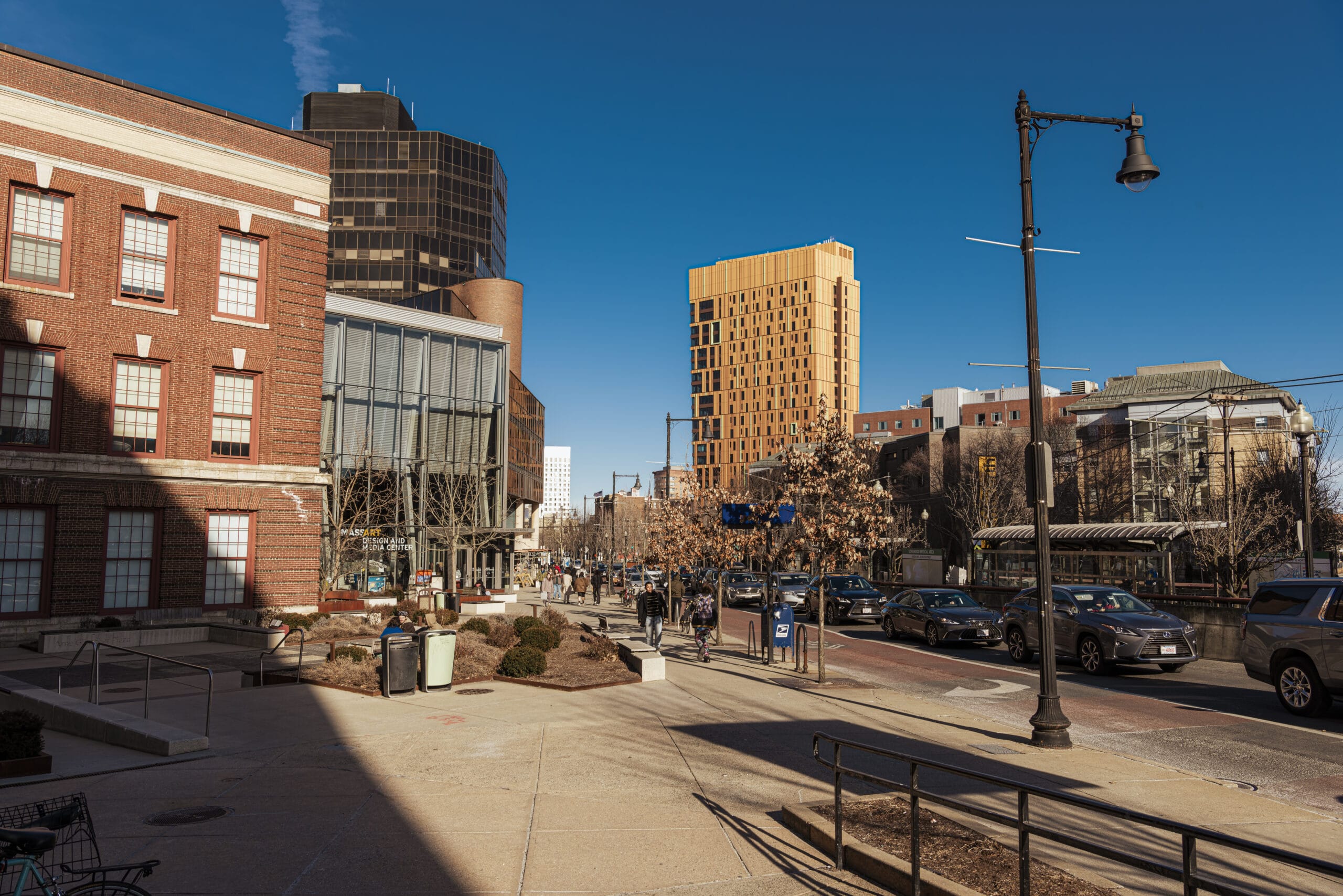 Urban street scene with a mix of modern and older brick buildings under a clear blue sky. Cars drive along the tree-lined street, where support resources for veterans are easily accessible. People walk on sidewalks, and a tall, rectangular orange building stands prominently in the background.