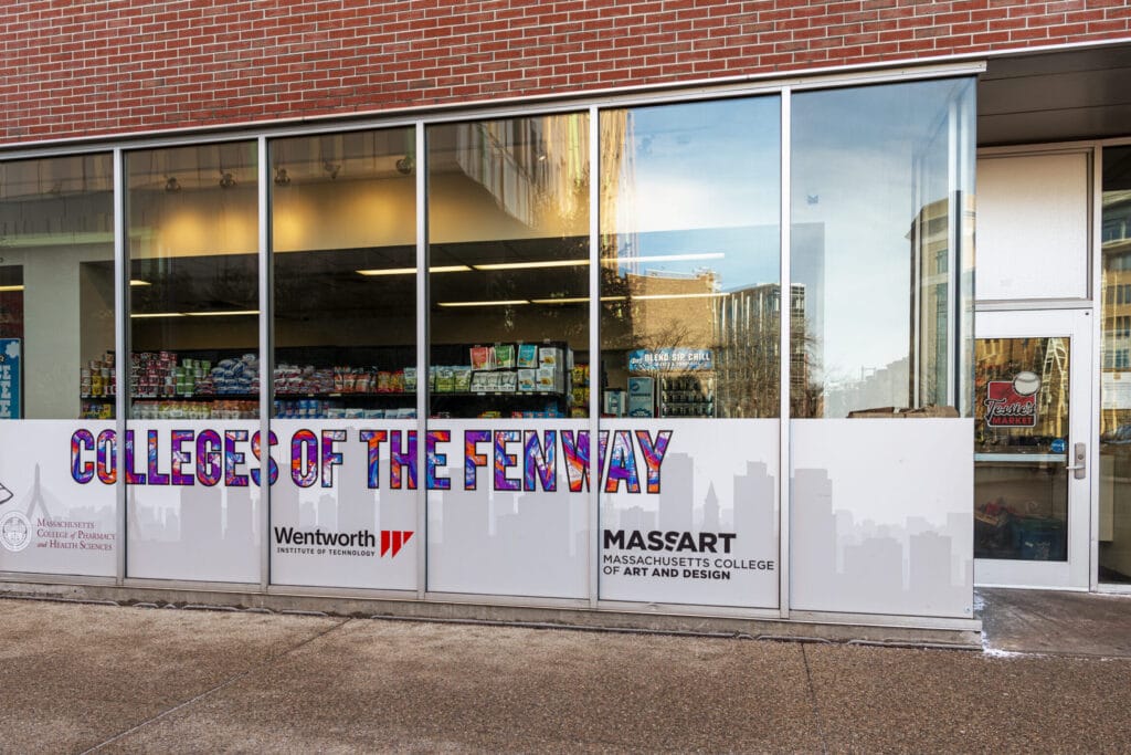 Storefront with large glass windows displaying College of the Fenway in colorful letters. Below, logos for Wentworth and MassArt are visible. Inside, shelves are stocked with dining essentials. Brick building and sidewalk in front offering a taste of housing life nearby.