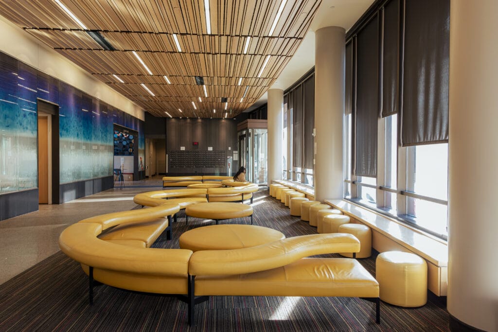 A brightly lit lobby with plush, curvy yellow couches, a wood paneled ceiling, and a blue mural on one wall.