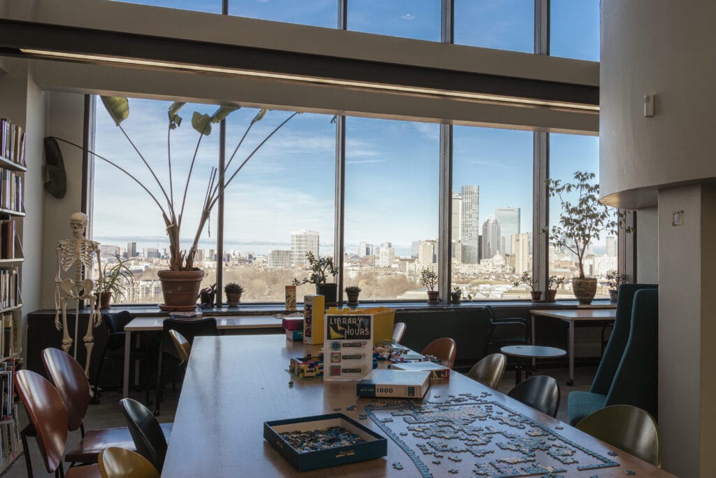 A modern library with large windows framing a city skyline. A puzzle is partially assembled on the table. Indoor plants and a skeleton model decorate the space, with shelves of books lining the walls. Natural light fills the room.