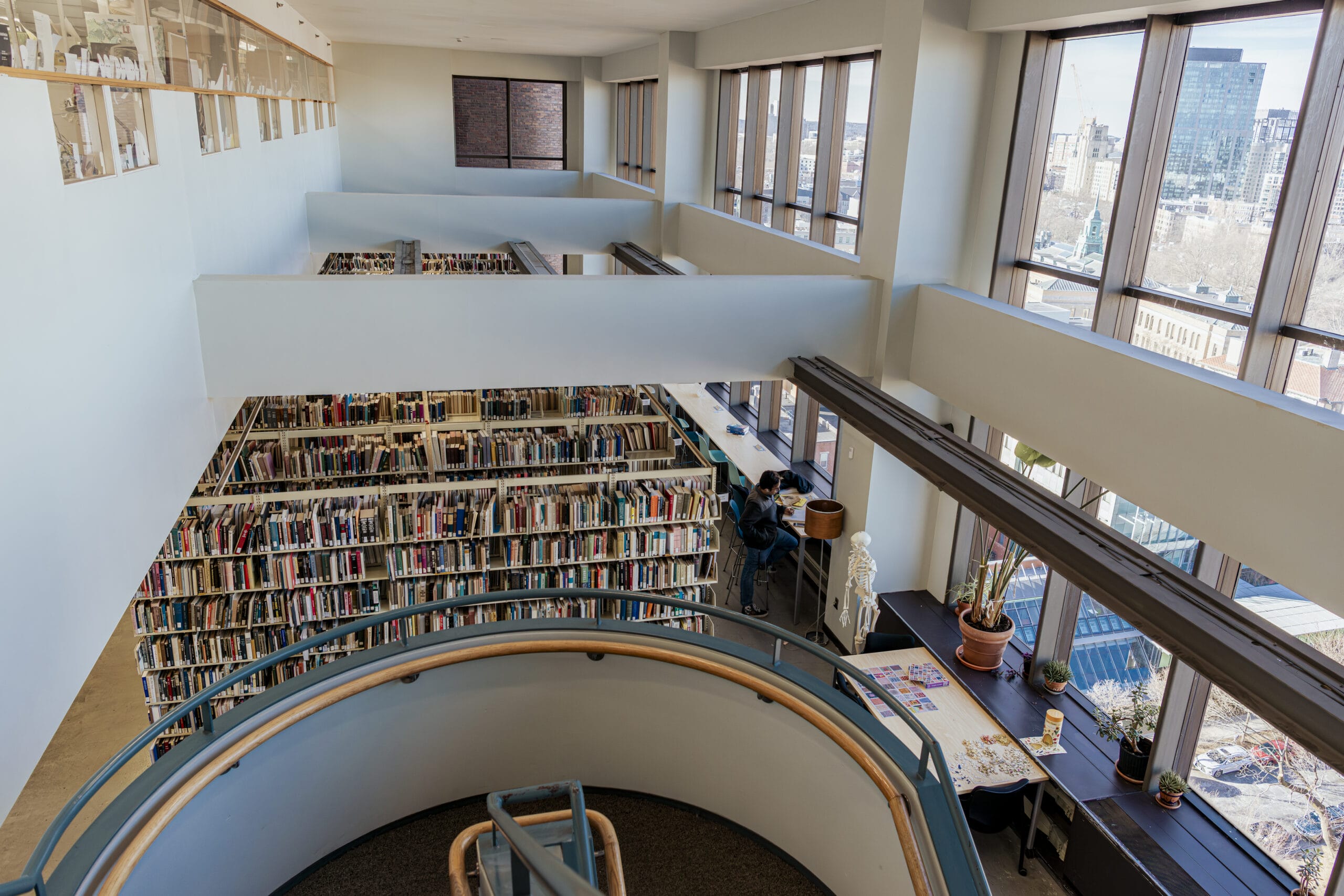 A multi-story library with large windows and rows of bookshelves. A person sits at a desk near the window, working on a laptop. There are potted plants and papers on the desk. A spiral staircase leads to the lower floor.