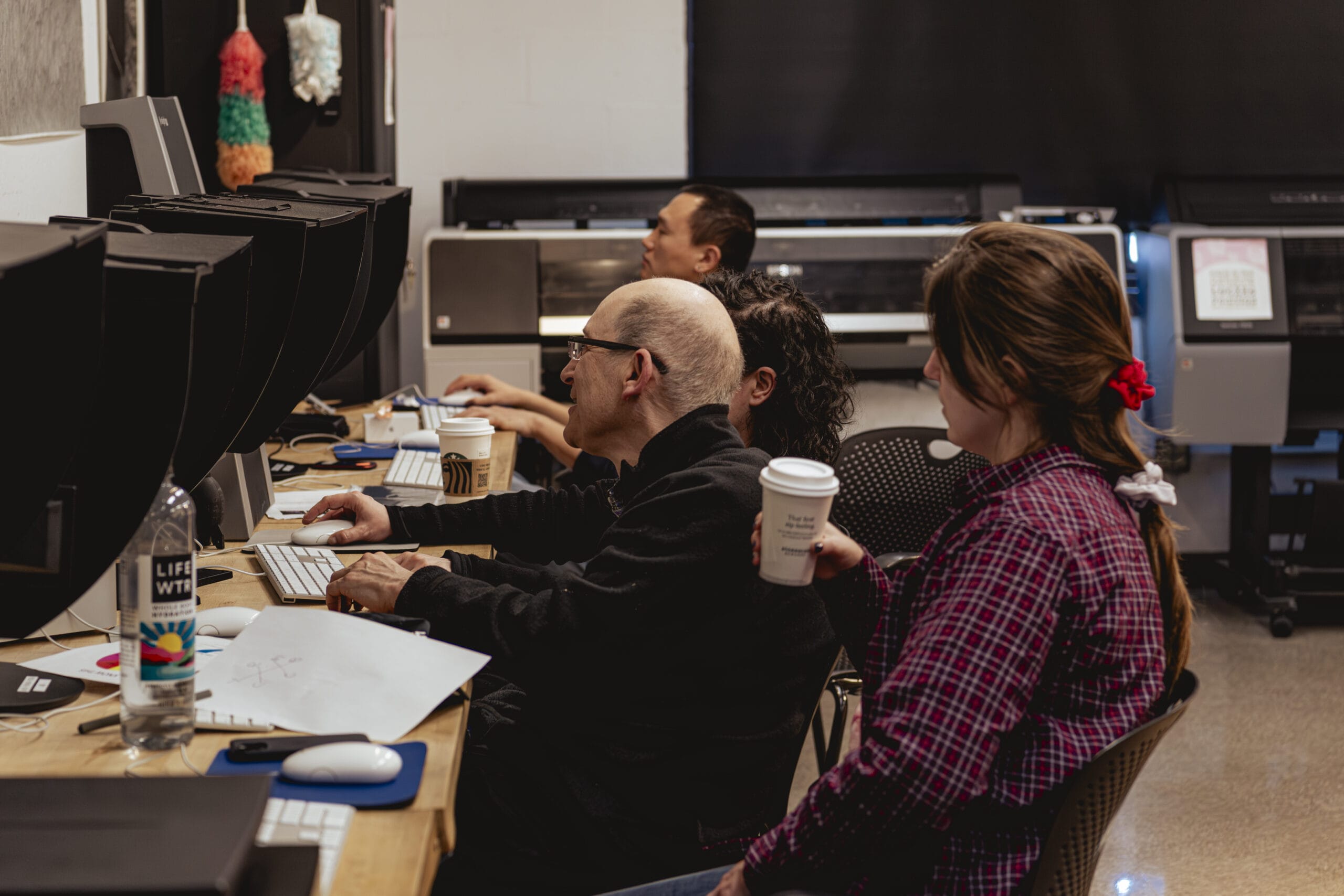 Four people are seated at a desk in a digital lab, working on computers surrounded by large printers. One person holds a coffee cup while another keeps a bottle of water nearby. A colorful piñata adds a touch of whimsy to the scene, reminiscent of creativity found in photography darkrooms.
