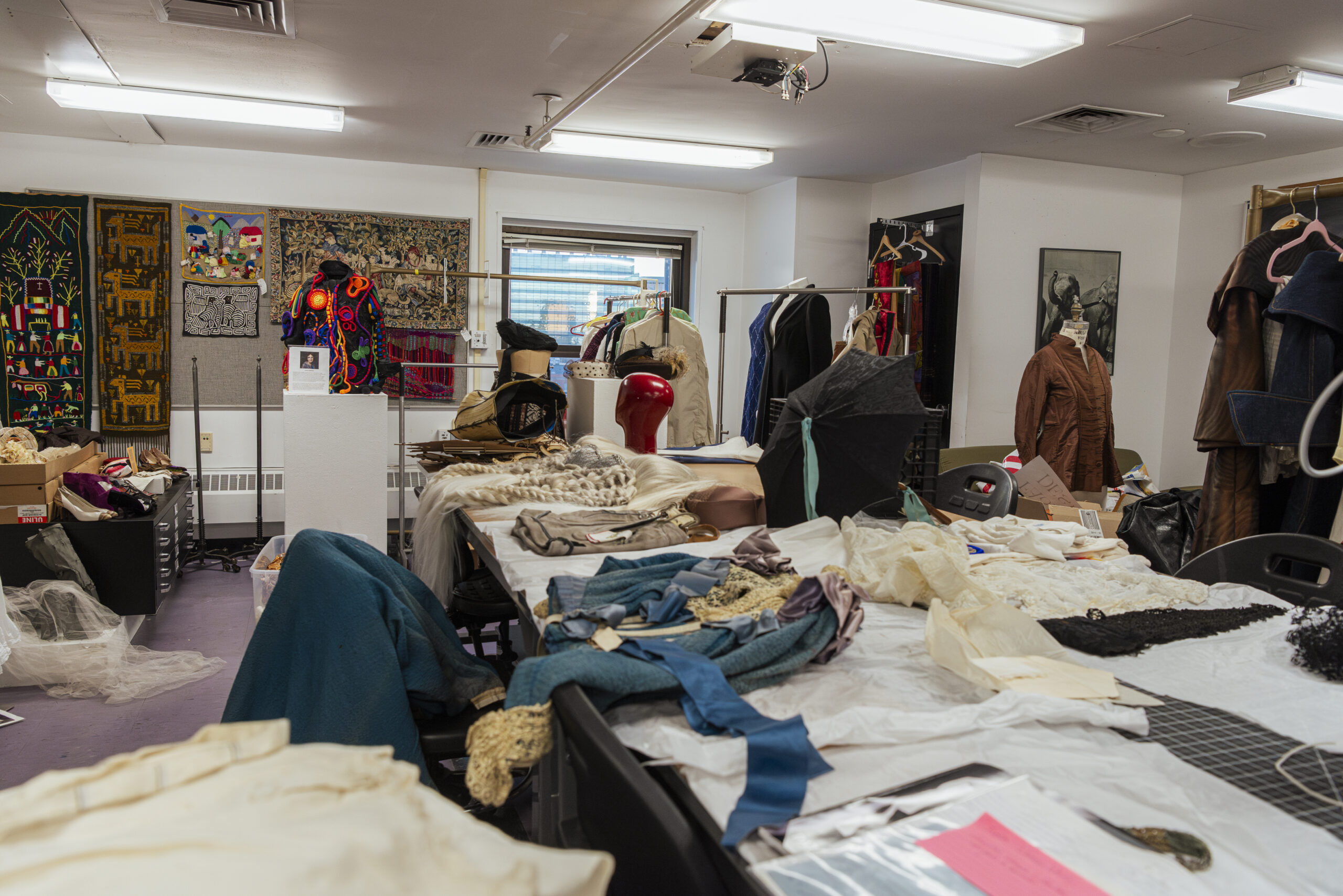 A cluttered workshop reminiscent of a fashion design studio, filled with various fabrics, vintage clothing, hats, and sewing materials. Garments and costumes adorn tables and mannequins, while colorful artwork decorates the walls, creating a vibrant and busy atmosphere.