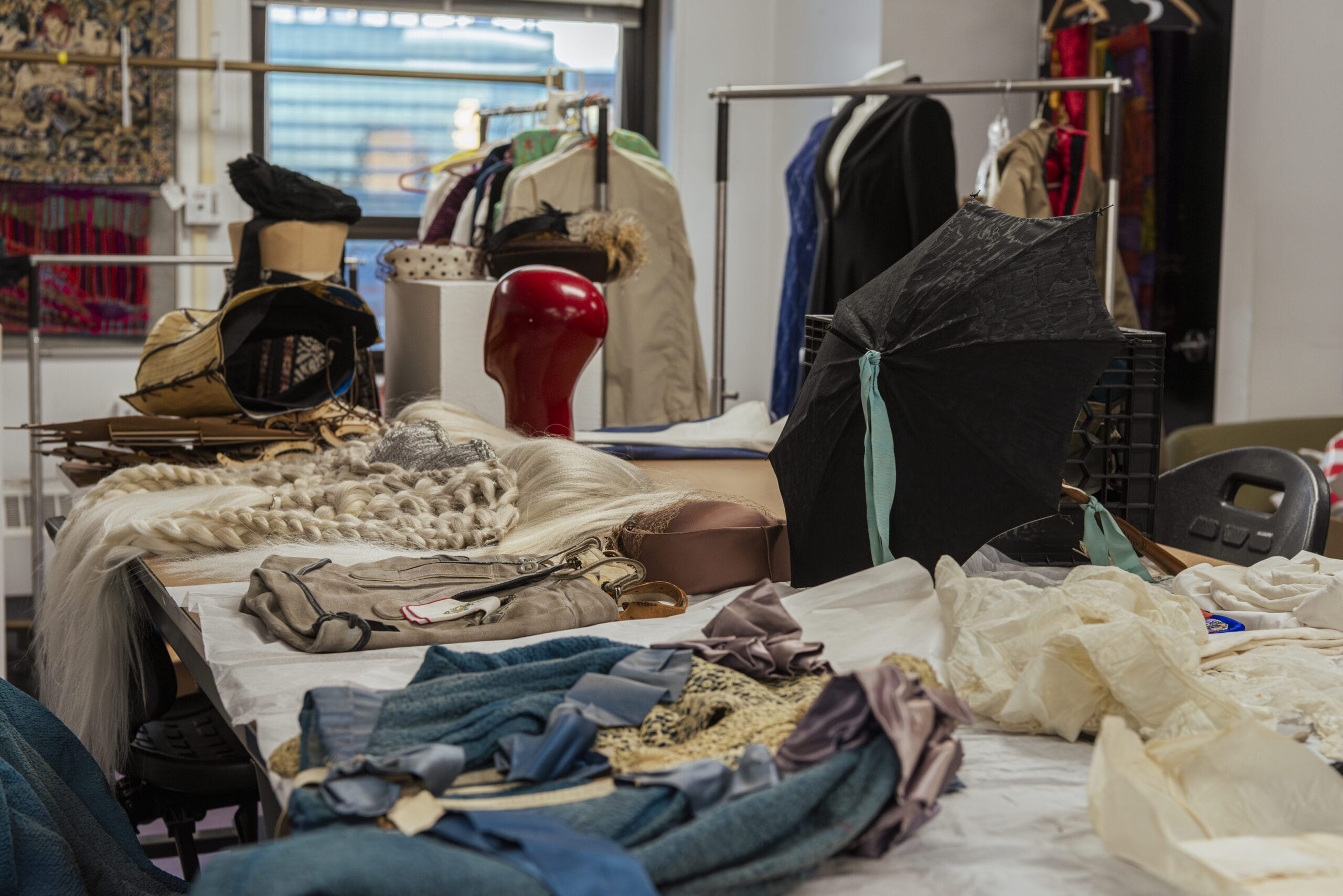 A cluttered fashion design studio brims with creativity: a table scattered with wigs, hats, an umbrella, and fabric pieces. Coats and dresses hang on a rack in the background, while a window reveals a glimpse of a building outside.