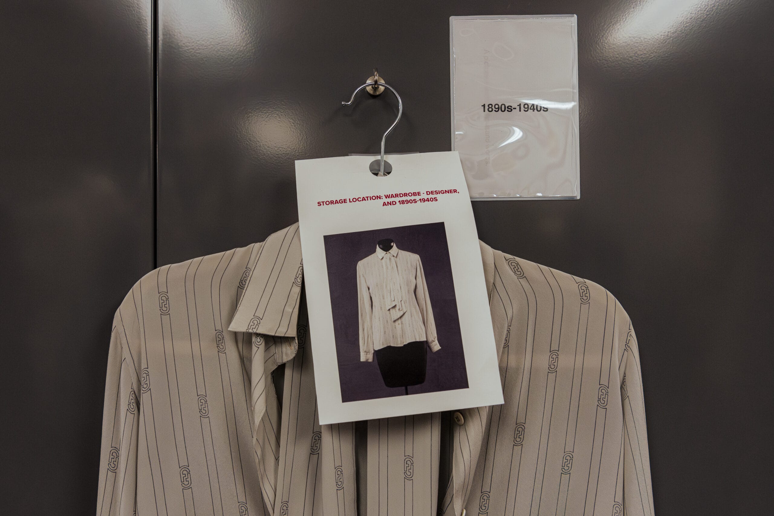 A striped beige blouse, styled with a creative touch, hangs on display in MassArt Fashion Design Studios. A card featuring its photo reads STORAGE LOCATION WARDROBE: RESOURCES AND 1980s-1990s, suspended against a textured dark surface labeled 1890s-1940s.