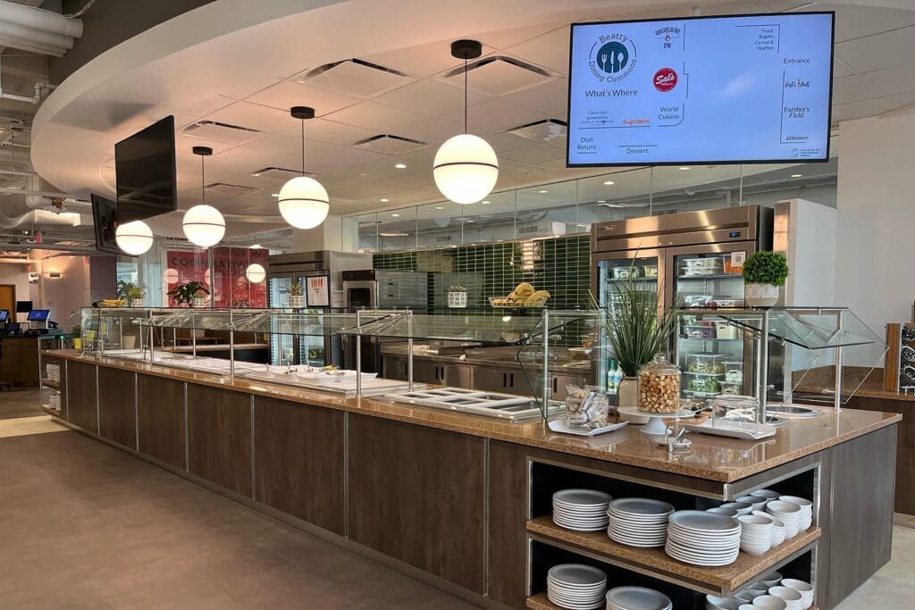 A modern cafeteria with a wooden counter and glass dividers offers a contemporary dining experience. Above, spherical pendant lights illuminate the space. Shelves display plates, and a digital menu is visible. The room has a clean design with green tiles and food displays.