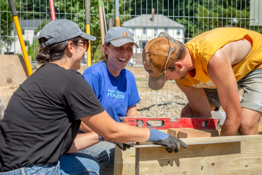 Three people collaborate on a community construction project outdoors. One person in a blue shirt smiles, another in black talks, and a third in yellow uses a level tool. Building materials are scattered about, showcasing their shared effort and camaraderie.