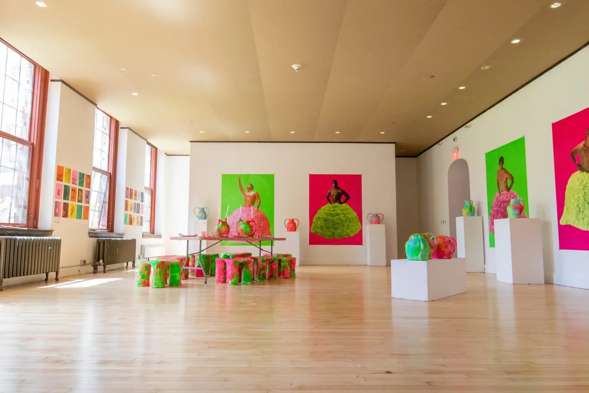 Godine Gallery is a bright art space with large windows, displaying colorful portraits of people in vibrant dresses, neon sculptures on pedestals, and various vivid artworks arranged along the walls and floor.