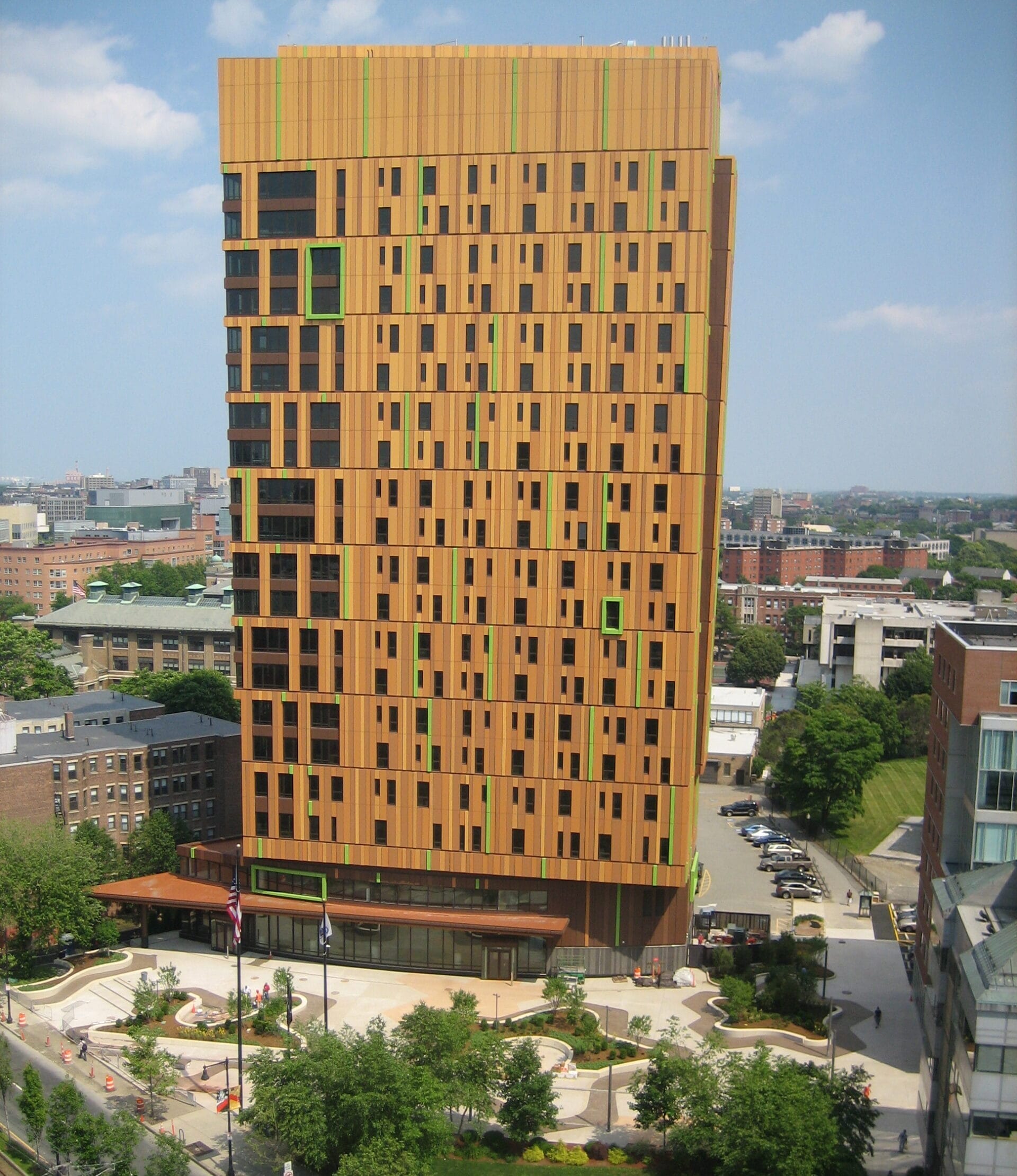 A tall, rectangular building with a unique orange and black patterned facade stands amid trees and buildings, offering vibrant on-campus housing. The structure features green accents against a blue sky with scattered clouds, while a landscaped area surrounds its base.