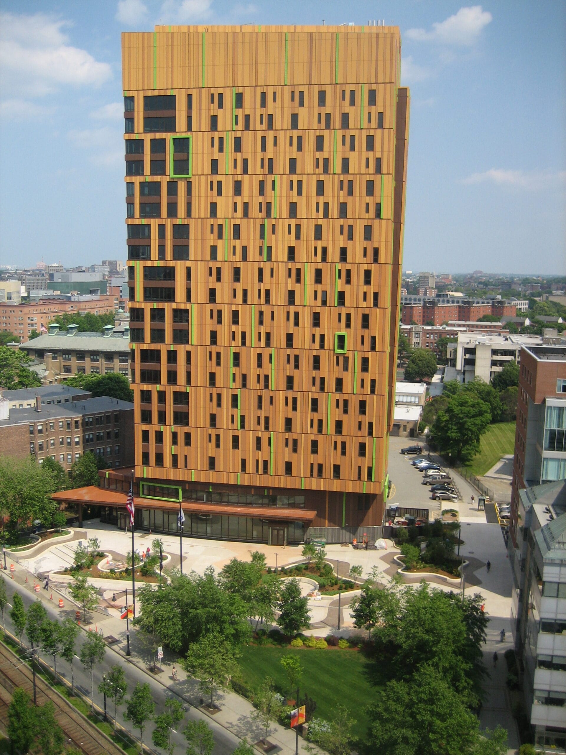 A tall, rectangular building with a unique orange and black patterned facade stands amid trees and buildings, offering vibrant on-campus housing. The structure features green accents against a blue sky with scattered clouds, while a landscaped area surrounds its base.