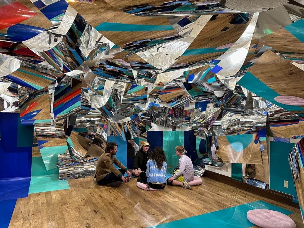 Four people sit on a wooden floor in a room with mirrored, geometric walls that reflect various colors, creating a kaleidoscopic effect akin to an MFA event. Blue and teal panels add contrast to the reflective surfaces.
