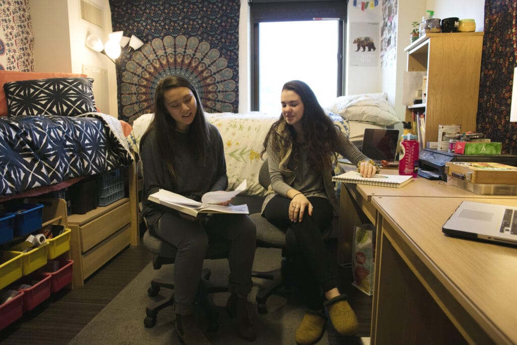 Two roomates sit together looking at a book with low bunkbeds in the background.