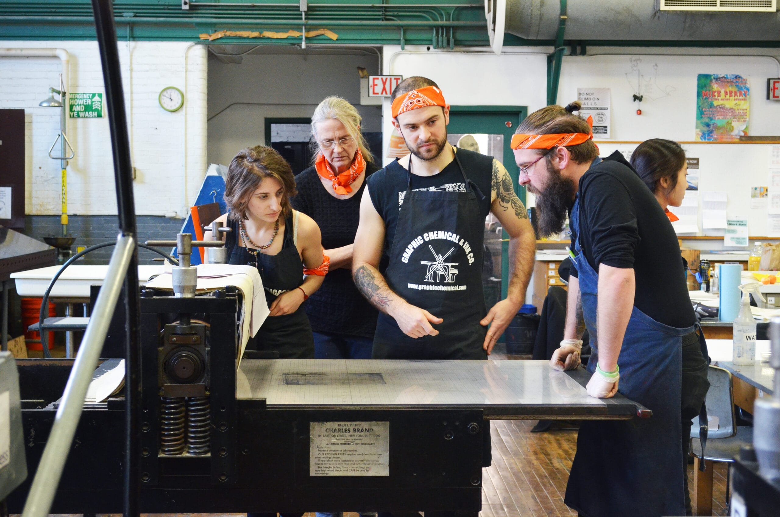 Students with orange bandanas stand around planning what they're going to print in the Print shop at MassArt.