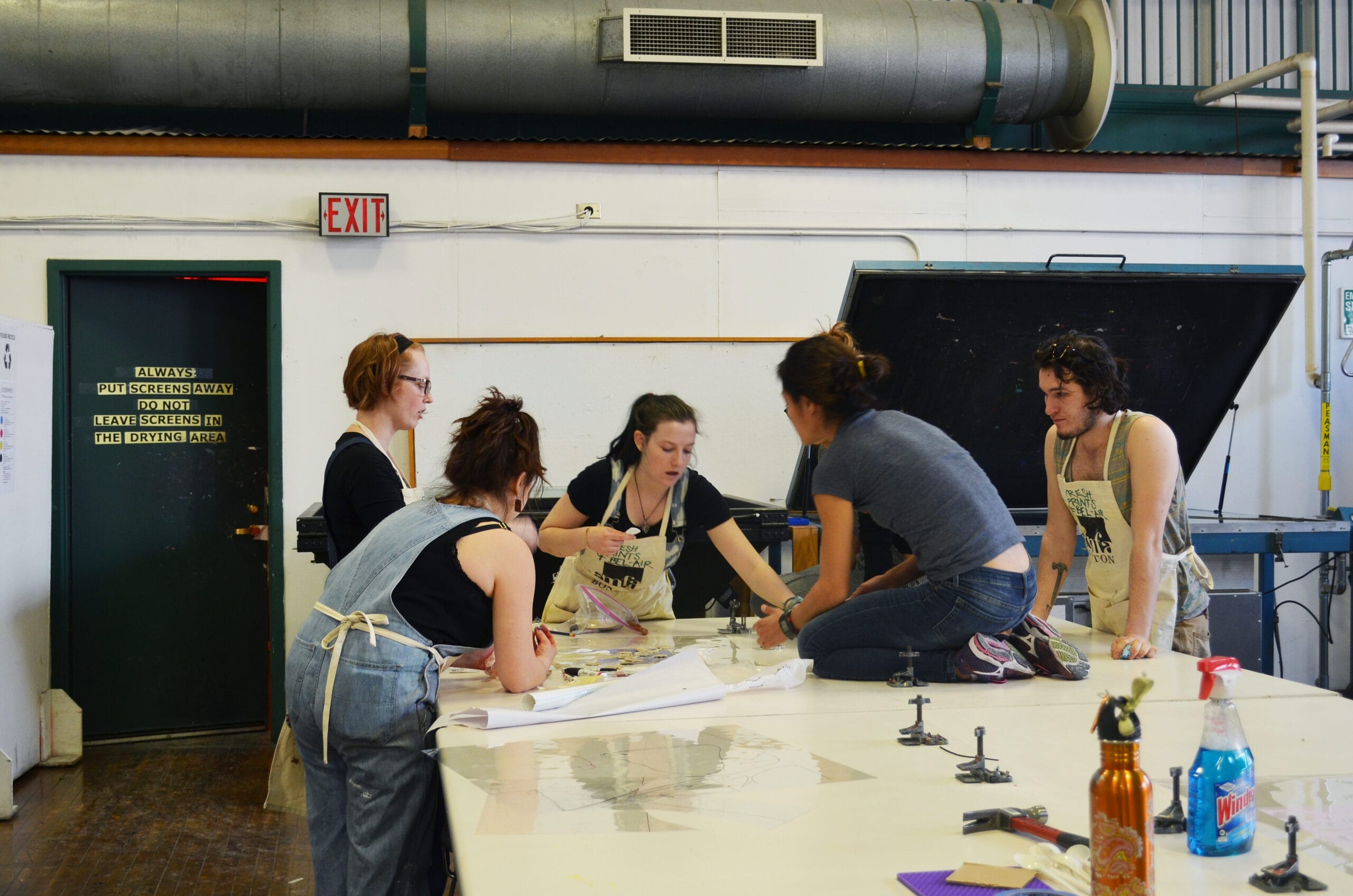 A group collaborates around a large table in an art studio, focused on a printmaking project. Tools and materials are spread across the surface, reflecting their creative endeavor. The space features industrial touches like a large vent and an exit sign, setting the perfect scene for printmaking events.