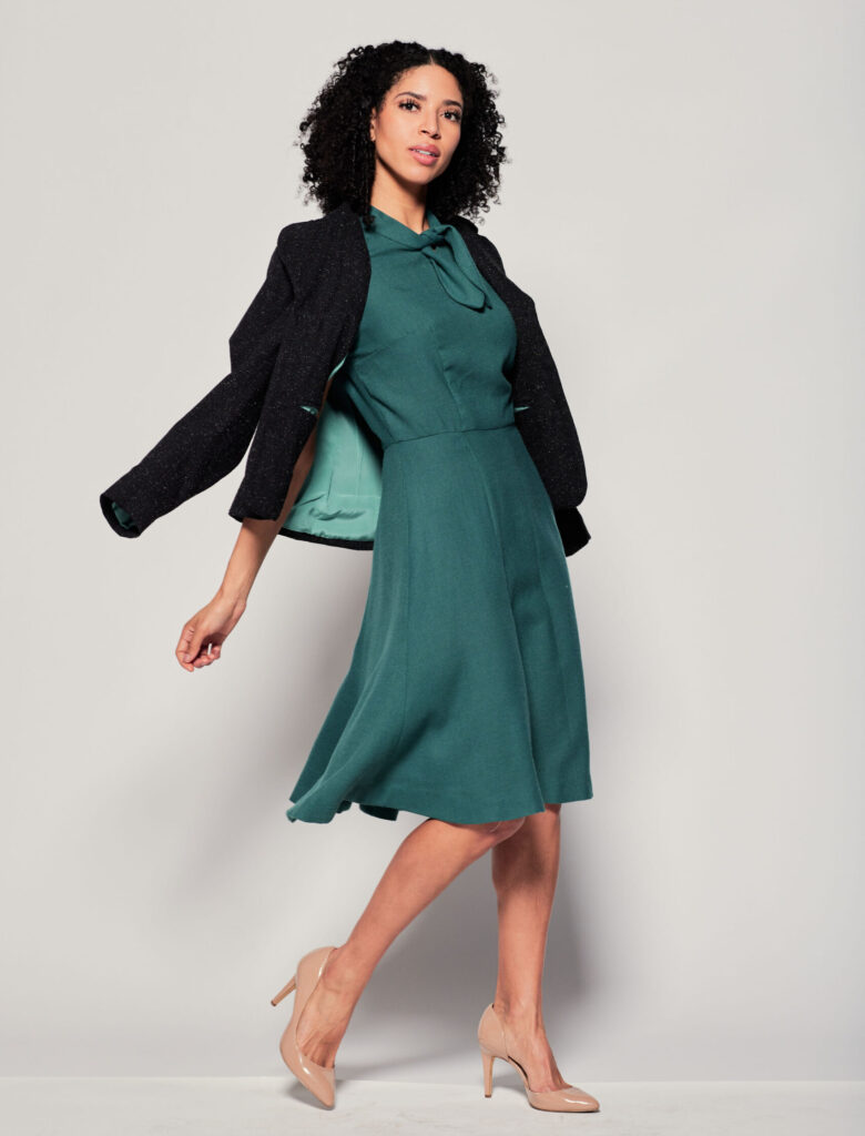 A model wearing a dark green dress with a black blazer thrown over her shoulders.