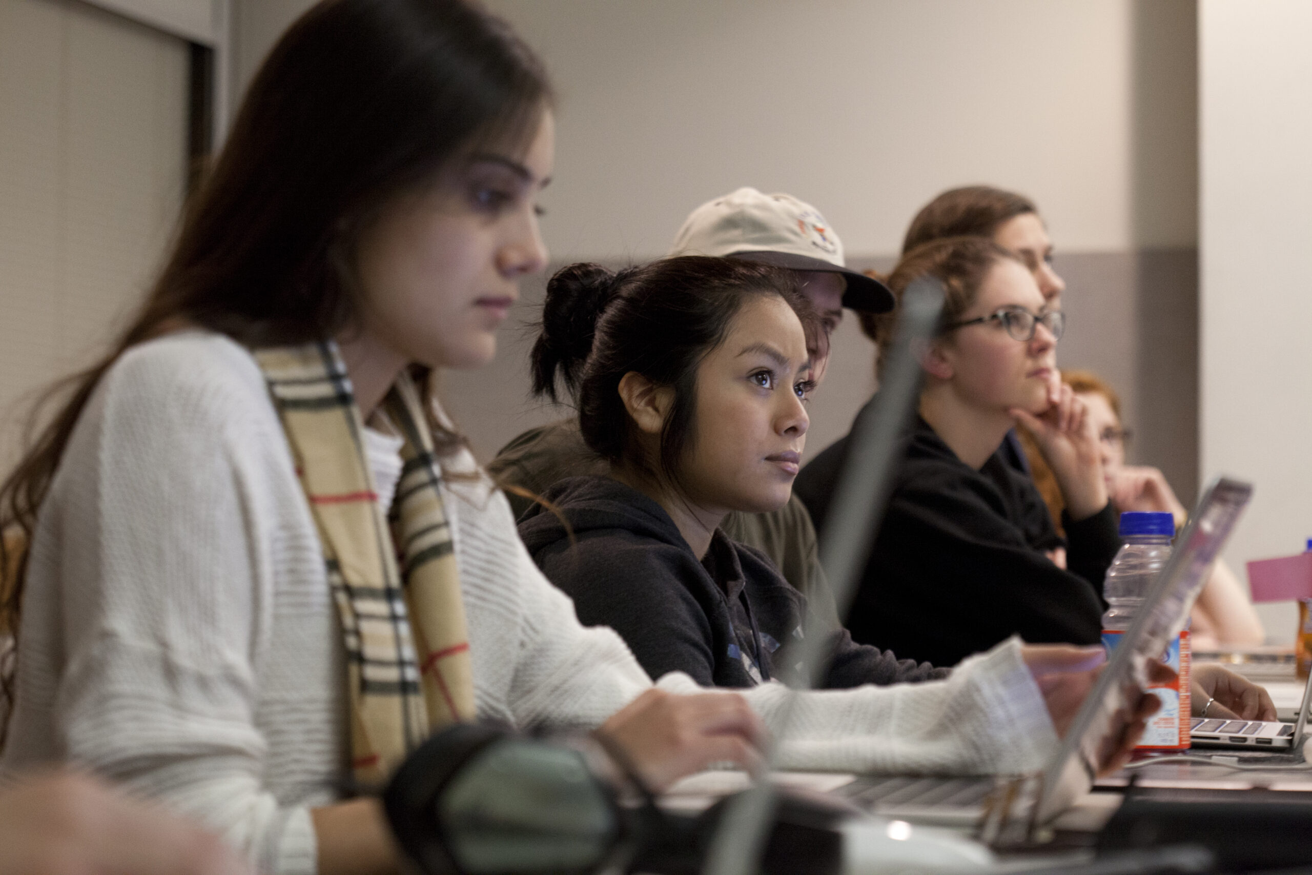 A group of focused MDes students sits in a well-lit classroom, attentively watching the front. They have laptops open, with some holding notebooks. A bottle of water is visible on the desk, underscoring their commitment to design innovation and an enriching learning environment.
