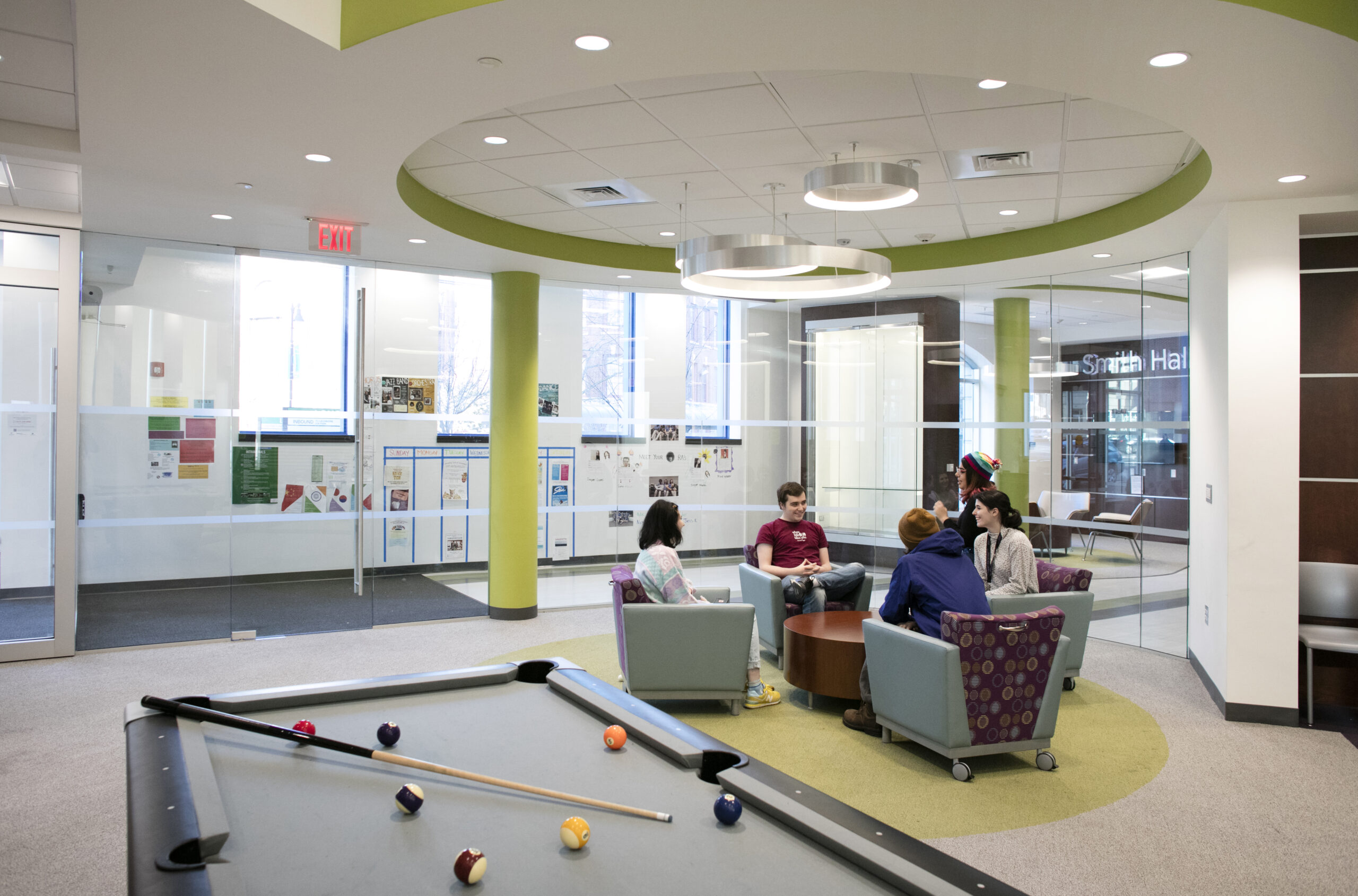 A pool table in the foreground with students sitting in chairs in a circle around a coffee table in the background.