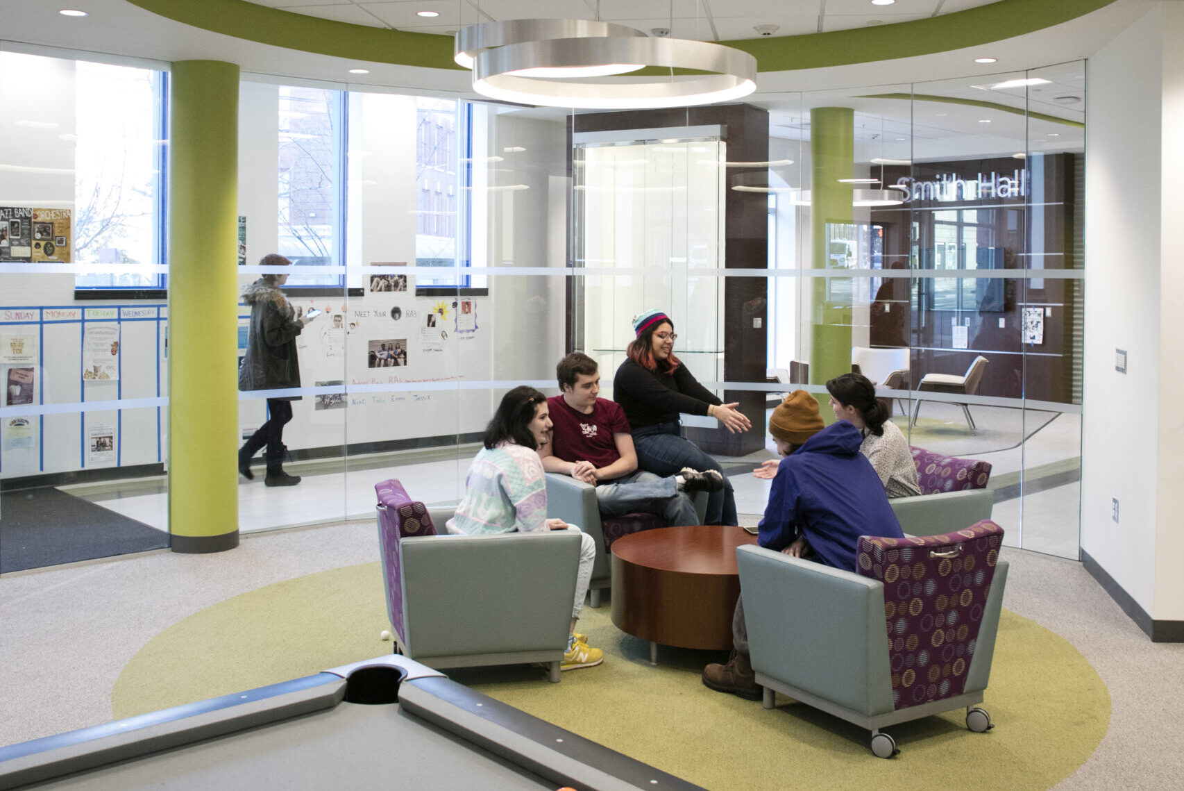 A pool table in the foreground with students sitting in chairs in a circle around a coffee table in the background.