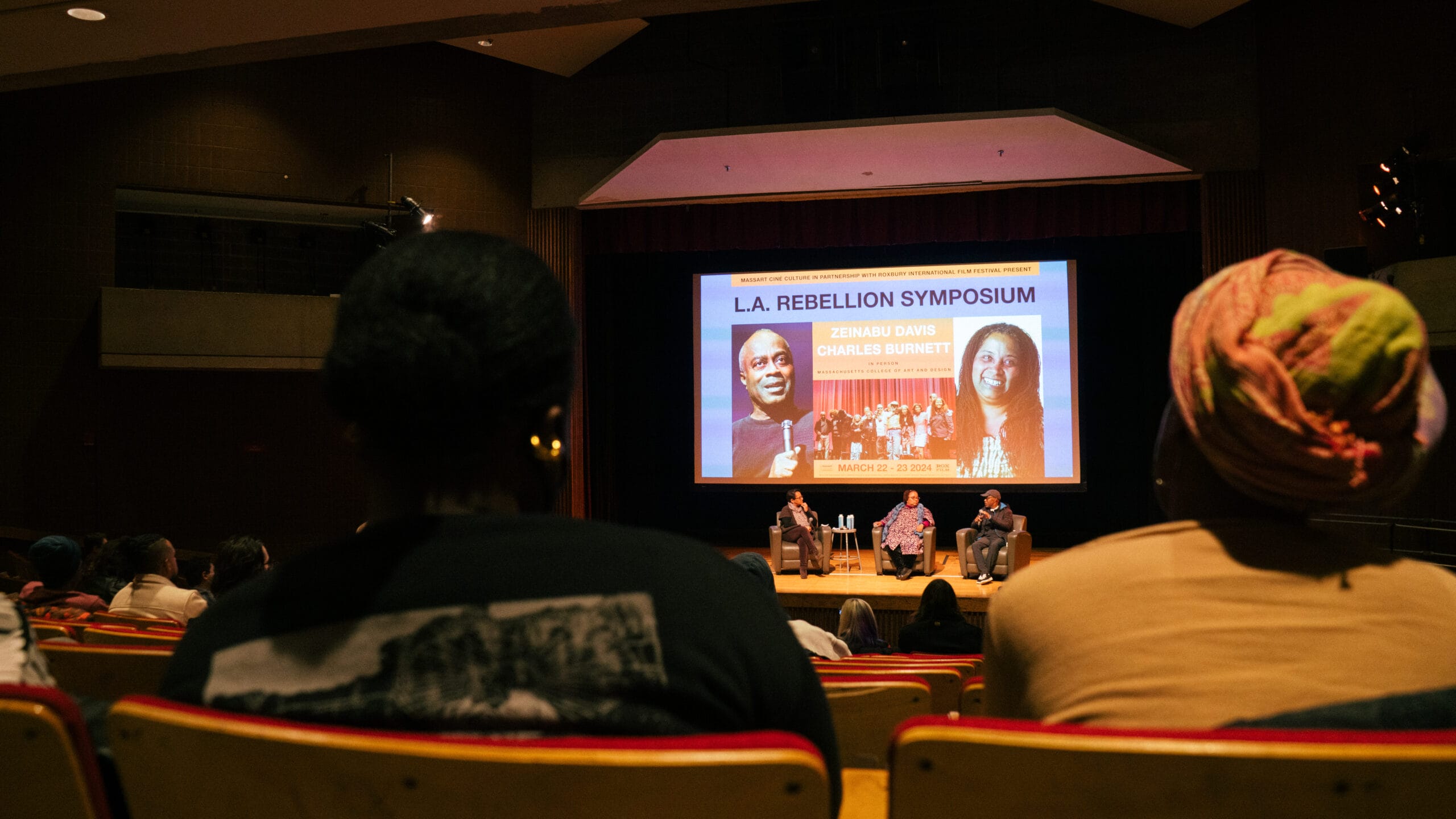 People sitting in an auditorium watching a panel of speakers with words "L.A. Rebellion Symposium" on the screen