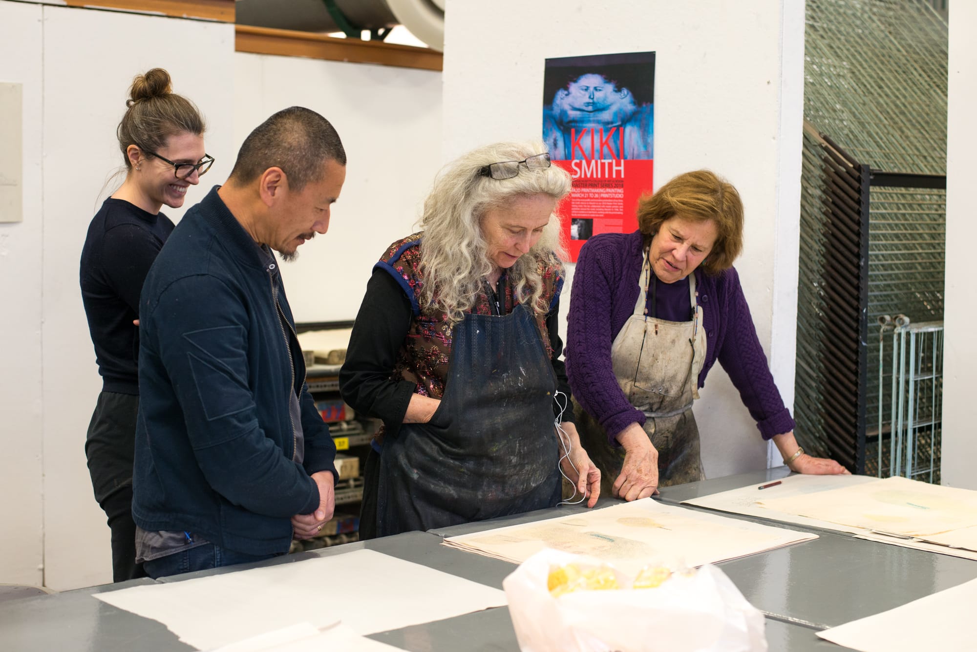 People wearing aprons stand around a metal table in a print studio, looking at prints.