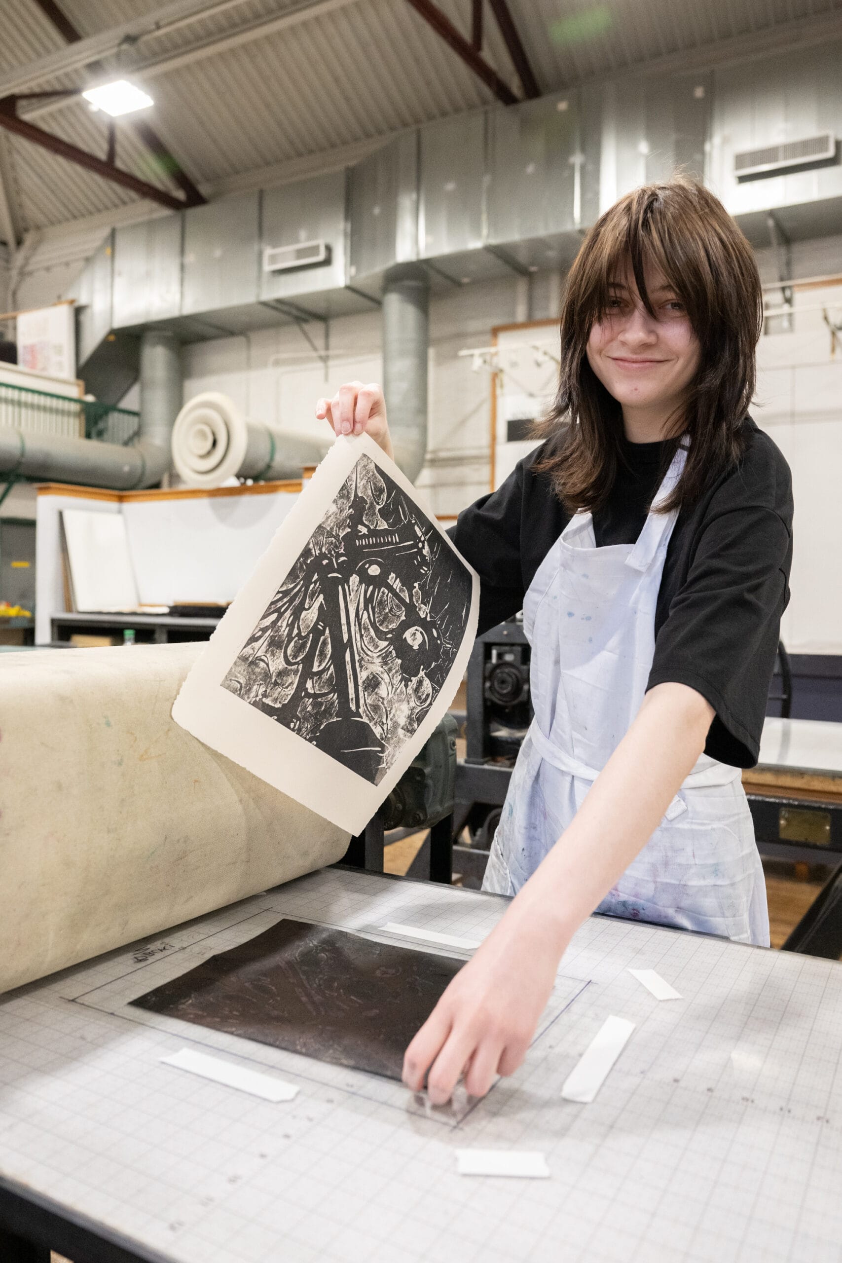A person wearing a black shirt and white apron smiles while lifting a freshly printed artwork from the press in a bustling printmaking studio. The art depicts an intricate design, ready to showcase at upcoming printmaking events. Industrial equipment is visible in the background.