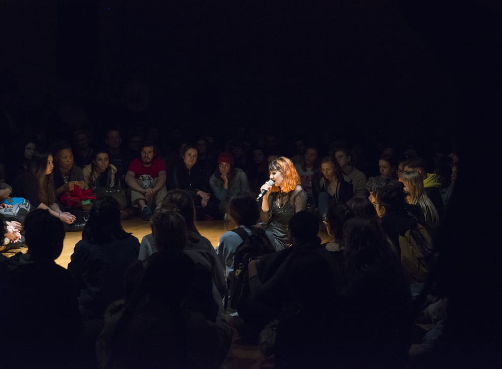 In the dimly lit room of SIM Studios, a performer with pink hair sits amidst a large seated crowd, microphone in hand. The audience is closely gathered around, creating an intimate atmosphere filled with anticipation and creativity.