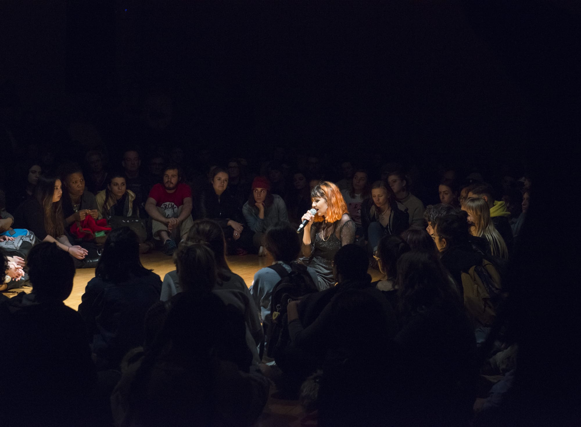 In the dimly lit room of SIM Studios, a performer with pink hair sits amidst a large seated crowd, microphone in hand. The audience is closely gathered around, creating an intimate atmosphere filled with anticipation and creativity.