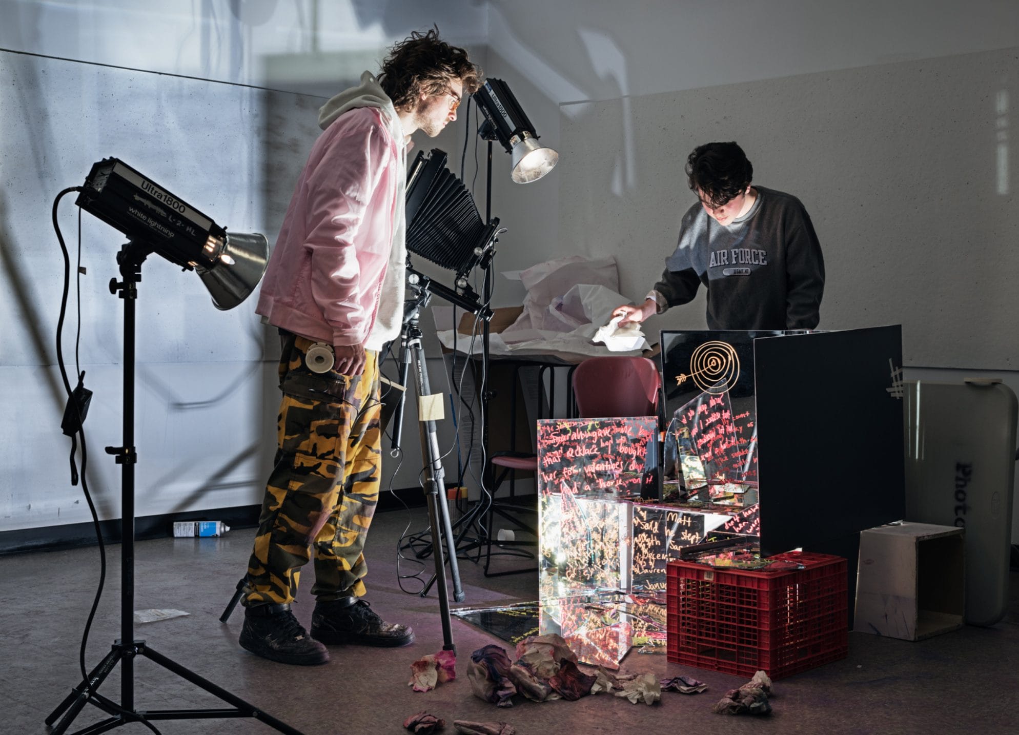 Two people work in a studio with bright lights, examining a table covered in colorful artwork and papers—an inspiring scene often found in a Photography BFA or Bachelor of Fine Arts program. One wears a pink jacket; the other, a dark sweatshirt, leans over the table.