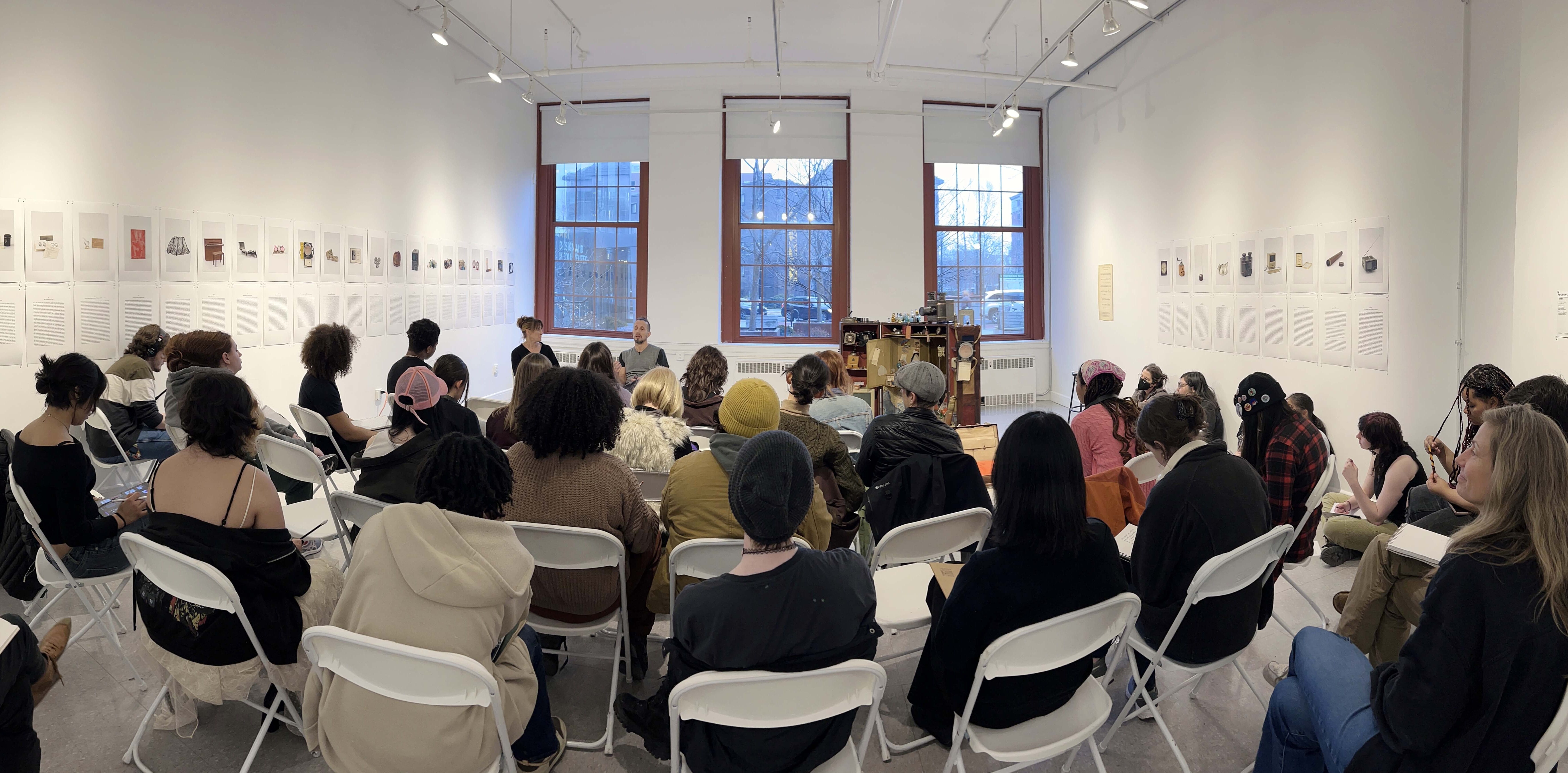 People seated on white chairs in Brant Gallery, facing speakers at the front; art hangs on the walls.