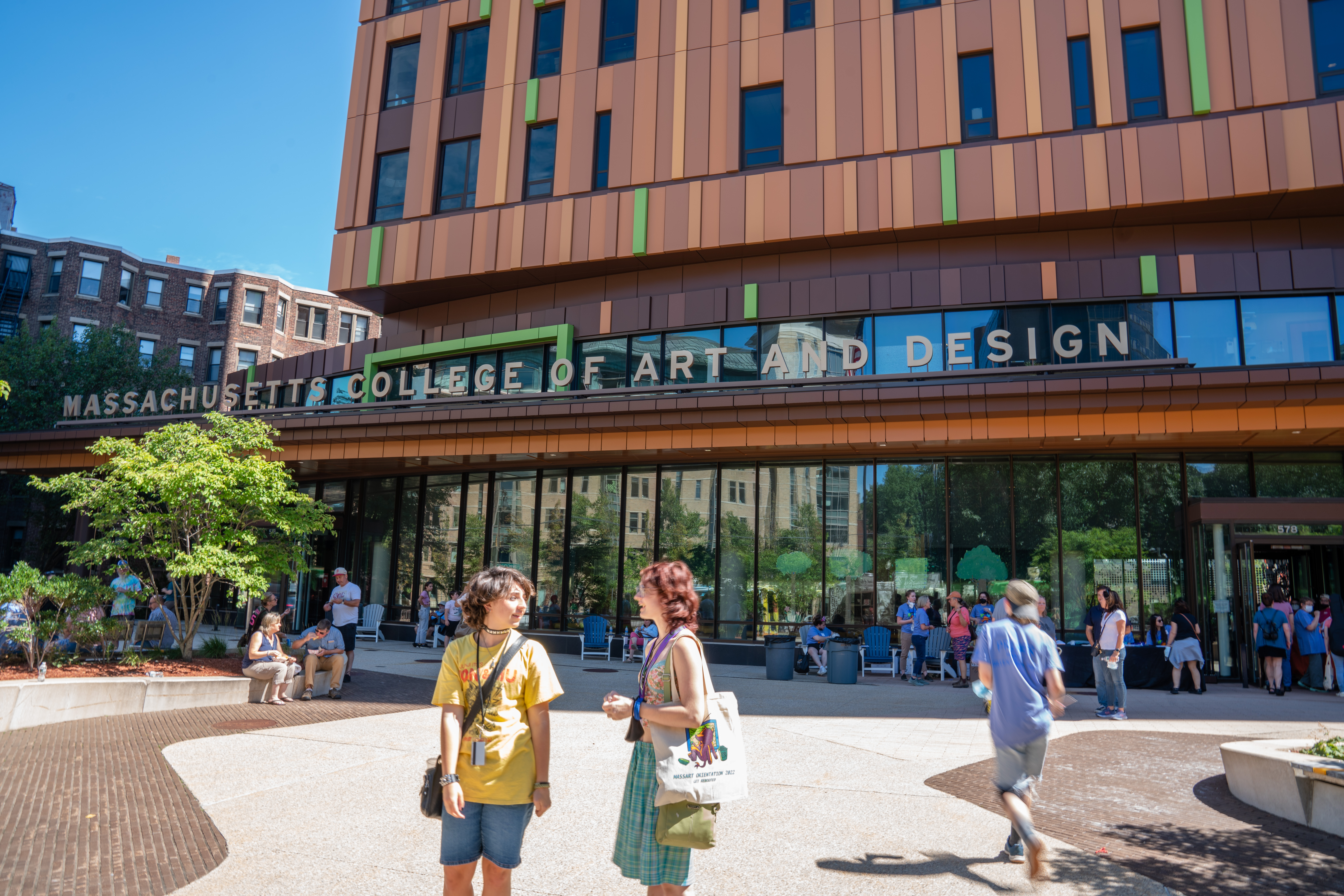 Students stand chatting in front of MassArt's Tree House Residence Hall as other students are seated and walking around in the background.