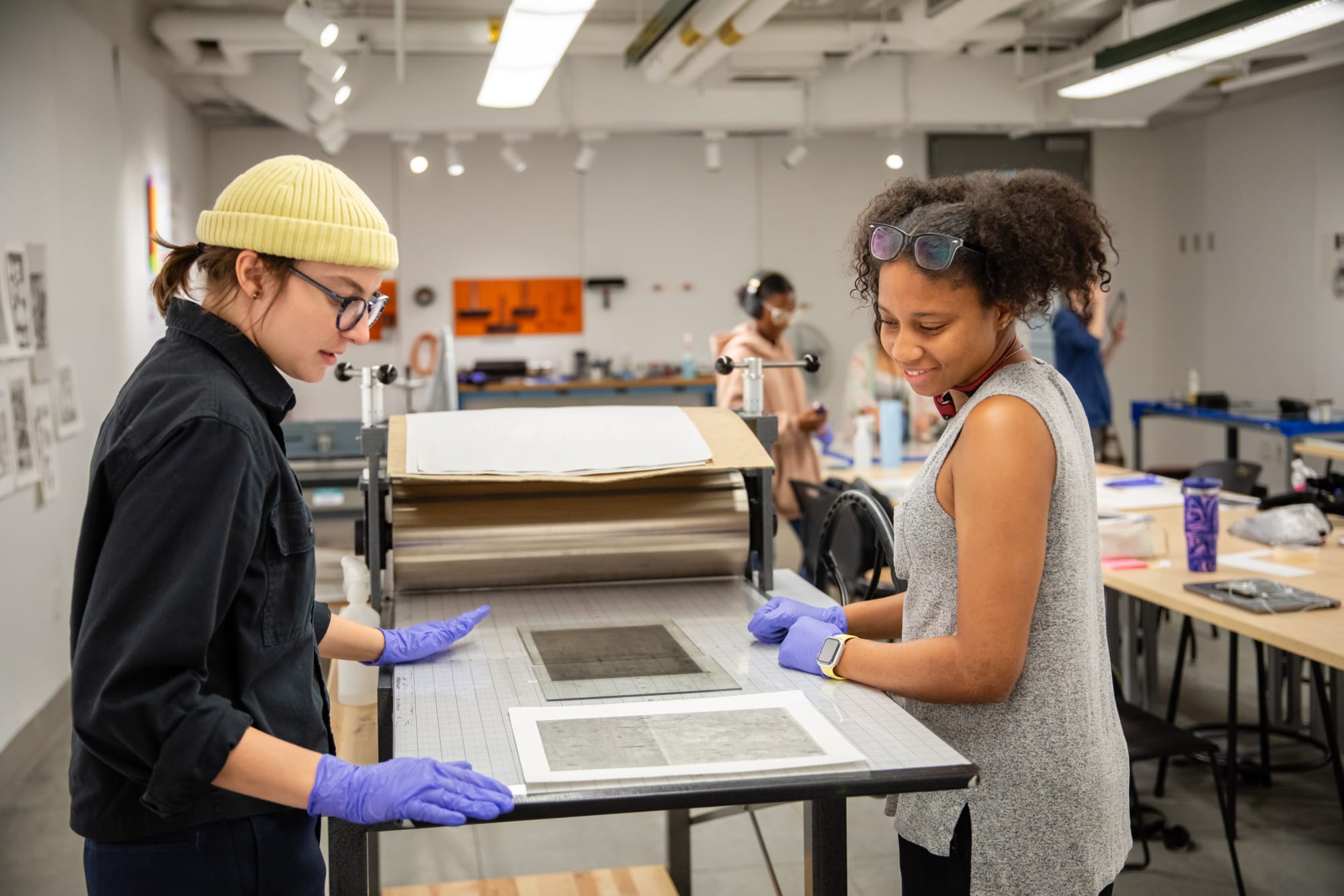 A woman in a wool hat stands in a printmaking studio talking to another girl looking at the print.