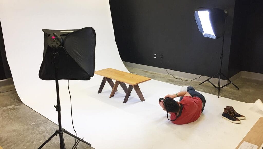 A person lays on the ground photographing a wooden bench in a room with studio lighting.