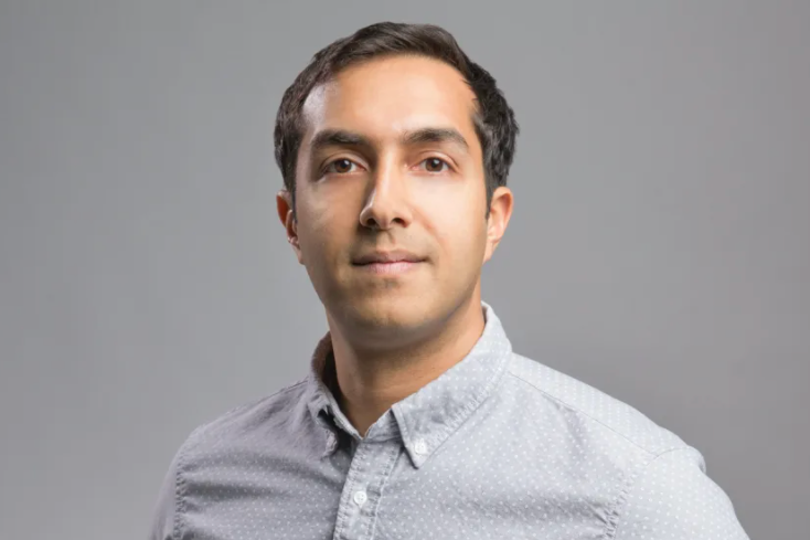 A person with short dark hair is wearing a light gray button-up shirt. They are standing against a plain gray background, looking confidently at the camera.