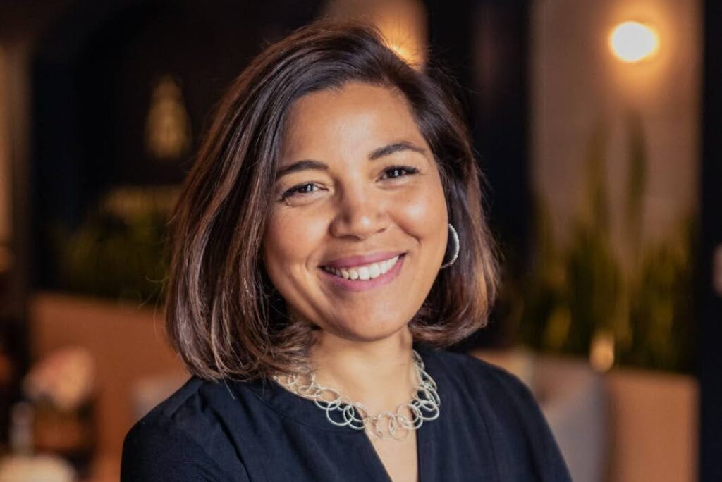 A woman with shoulder-length hair stands indoors, smiling with her arms crossed. She wears a dark blouse, a silver necklace, and a bracelet. The background features blurred plants and modern decor.