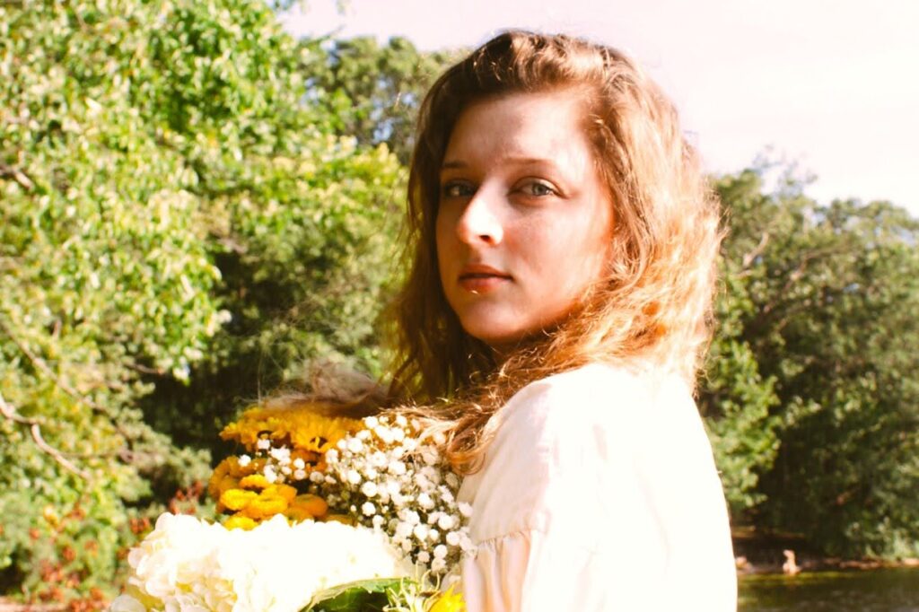 A woman with wavy hair stands by a lake, holding a bouquet of sunflowers and other white flowers. She wears a light-colored dress and looks over her shoulder. Lush green trees are in the background, and the lake reflects the sky.