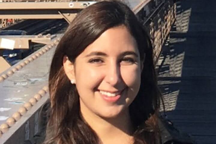 Woman smiling on a sunny day, standing on a bridge with metal railings. She has long brown hair and is wearing a black coat. The background shows architectural details and a glimpse of the sky.