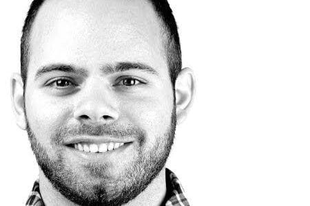 Portrait of a smiling man with a short beard and short hair, wearing a checkered shirt. The background is plain white, and the image is in black and white.