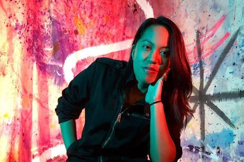 A person wearing a dark jacket sits on a stool, resting their chin on one hand. The background is a vibrant, colorful abstract mural with splashes of pink, blue, and orange hues.