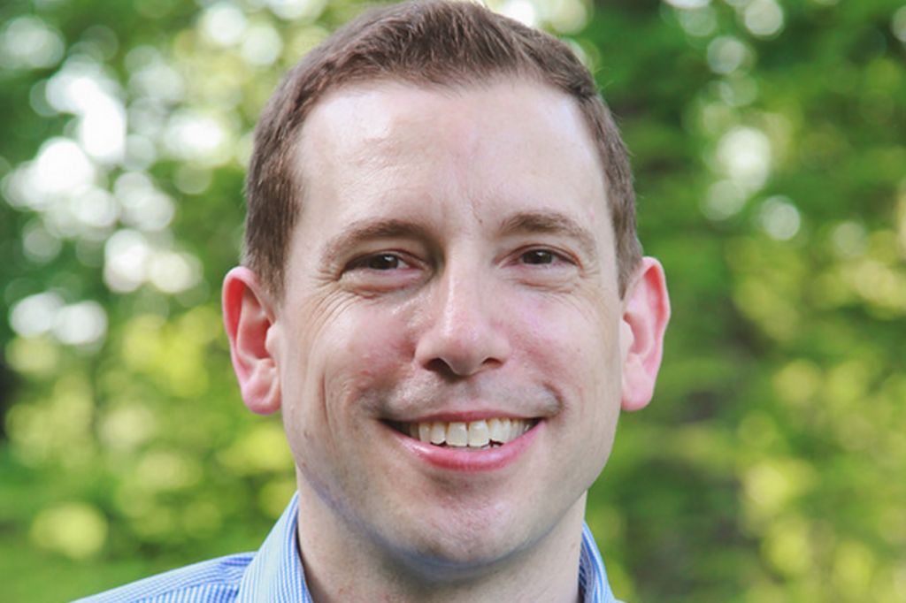 A person with short brown hair is smiling outdoors. They are wearing a blue, checkered button-up shirt. The background features blurred green foliage, suggesting a natural setting.