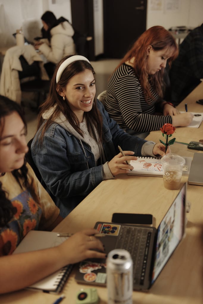 A group of young adults sits at a table with laptops and notebooks, immersed in writing and drawing. A smiling woman in a denim jacket holds a pencil and a notebook adorned with a red rose. Drinks, stickers, and resources for accessible education decorate the vibrant workspace.