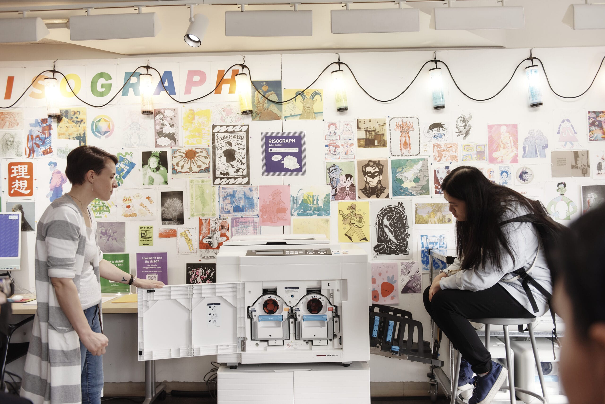 Two people are in a creative studio lined with colorful art prints. One stands by an open printer, showing its inside, while the other sits on a stool nearby. The space is bright with string lights above.